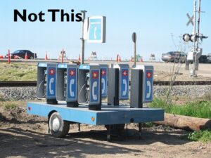 An image of several portable pay phones mounted ontop of wheel trailer that are in inaccessible reach ranges with the text "Not This" at the top of the image.