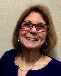 Marcie Roth, a white woman with shoulder-length light brown hair and red glasses, smiles at the camera. She is wearing a blue blouse and a pearl necklace.