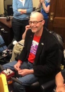 Mike Oxford, a bald white man with glasses, smiles at the camera while sitting in his motorized scooter. He's wearing a red graphic t-shirt and black blazer.