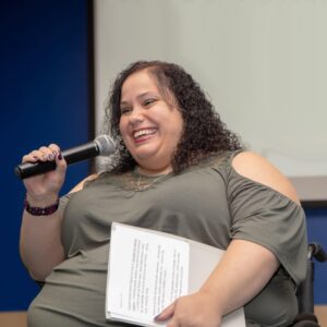 Millie Gonsalez, a light-complexioned woman with dark brown curly hair, smiles at something off camera while presenting.