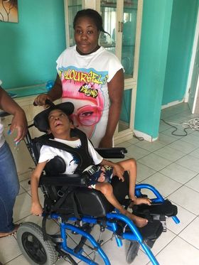 Devon, a young disabled black man, sits in in his first wheelchair, a manual chair with a blue frame. A woman relative stands nearby smiling.