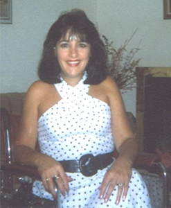 Photo of Benny, smiling, long black hair, wearing white dress with black polka dot dress, seated in her wheelchair.