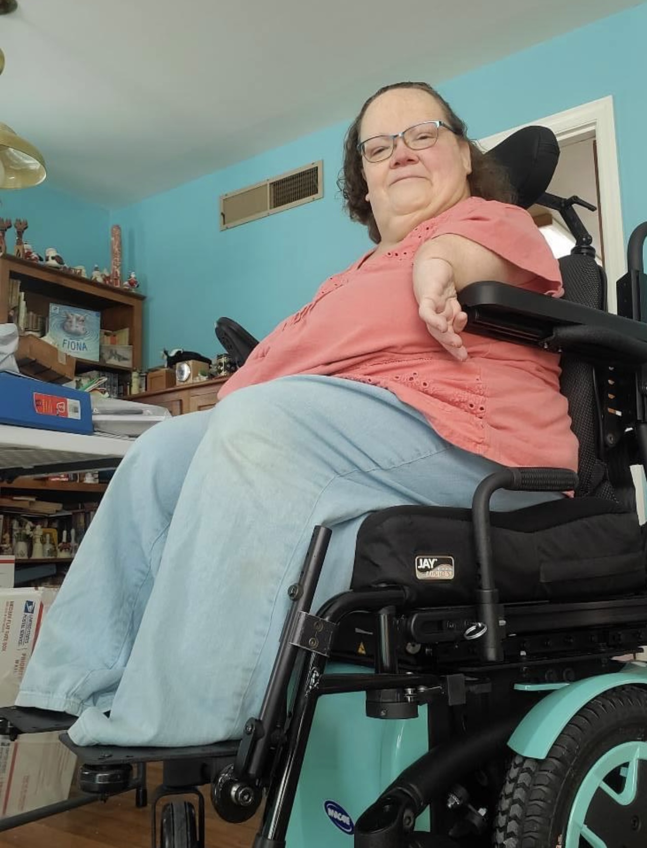 Gwen, with foreshortened arms, waering jeans and a peach-colored shirt, smiling and sitting in her power chair.