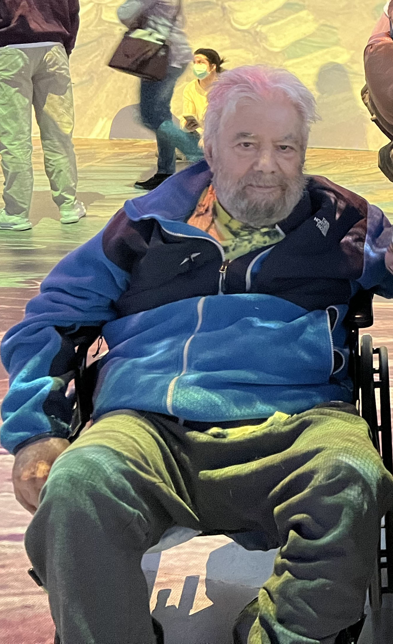 Photo of Keith using a wheelchair. He is an older man with white hair and a beard; he smiles at the camera. He is wearing a winter jacket and sweatpants.