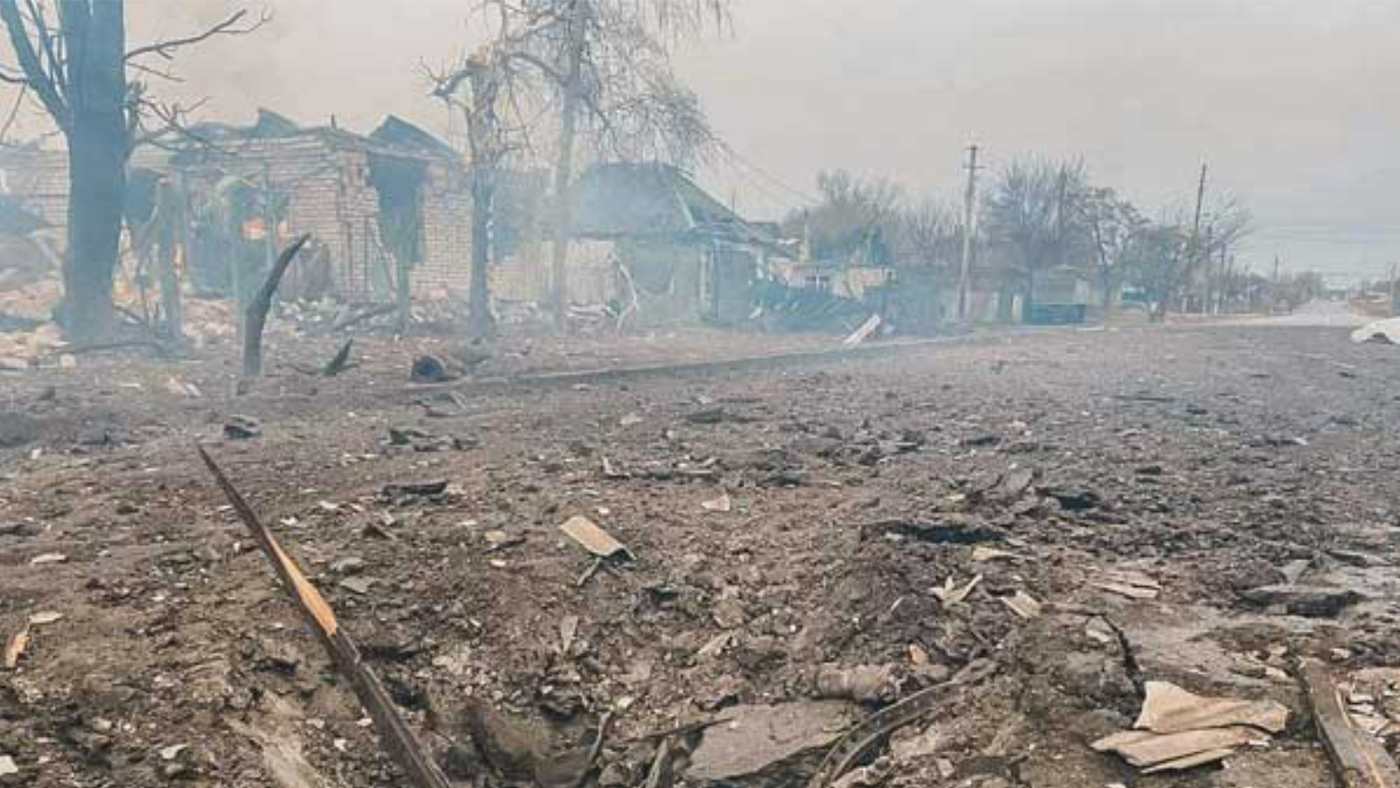 An utterly devasted suburban area in Ukraine; debris litters the street, and homes have been leveled. It is smiky and hazy, difficult to see into the distance.