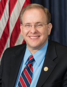 Photo of Jim Langevin in a suit and red, white and blue tie, in front of an American flag. He is a white middle-aged man with blond hair and glasses, smiling.