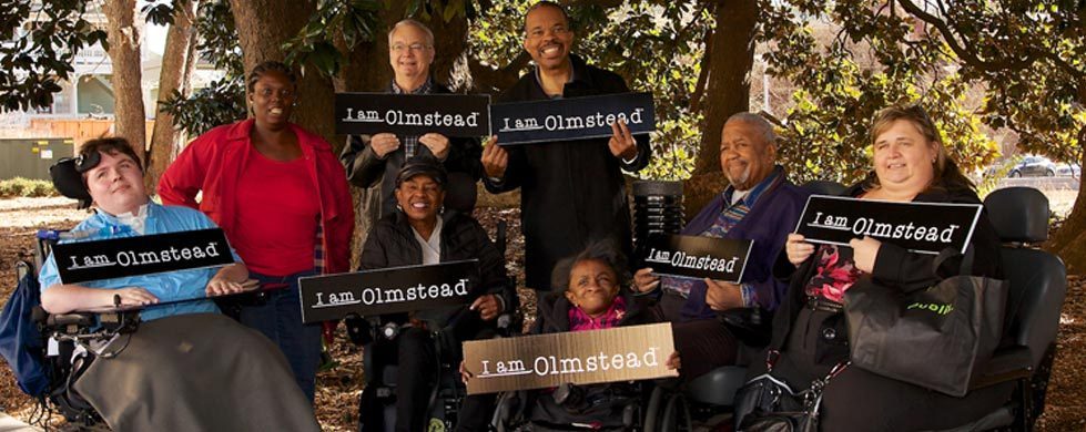 Image description: Group photo shot outdoors under a group of trees on a sunny day, from https://www.olmsteadrights.org/. The image includes 8 people of different colors and ethnicities, 5 of whom use wheelchairs, all smiling and holding signs that say “I am Olmstead.”