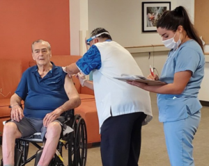 A white older man in a wheelchair receives aa vaccine from a man in scrubs; a woman in scrubs holds a clipboard and looks on. The medical professionals are wearing masks.