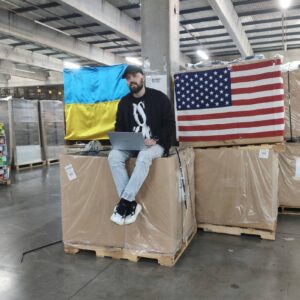 Image of FFR staff sitting on the packages in the warehouse in Ukraine the Ukraine and USA flag are behind him.