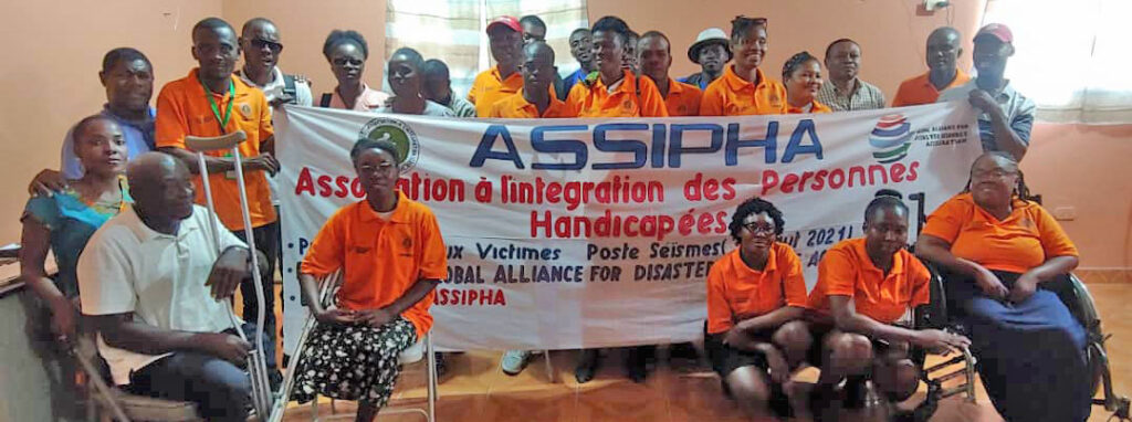 Un grupo de personas con diversas discapacidades, la mayoría con camisetas naranjas, todes de piel oscura, sonriendo y sosteniendo una pancarta blanca que lee: The Association à I’Intégration des Personnes Handicapées (ASSIPHA) y contiene también el logo de GADRA.