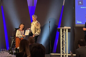Photo of Anna receiving her Top 4 award. She is wearing a white long sleeve top with dark orange pants; She is smiling, seated in her wheelchair, standing next to a woman who is holding a plaque between them. They are both on a dramatically lit stage.