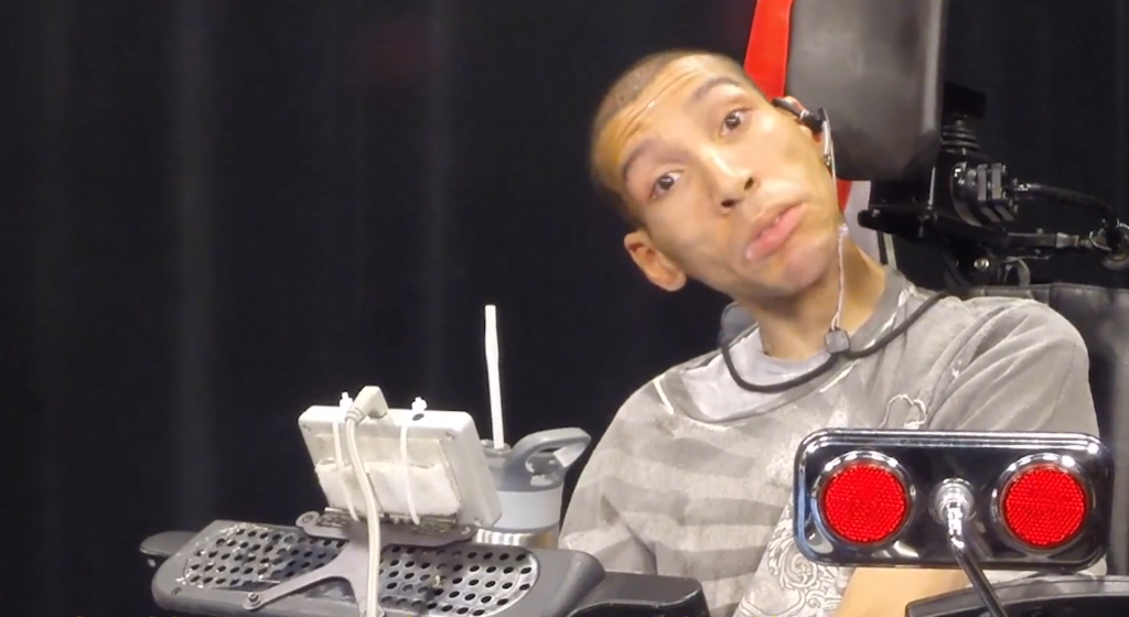 Flip, a Latino man, sits in his wheelchair with his communication device in front of him, looking at the camera with a serious expression.