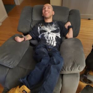 Flip, a Latino man with cerebral palsy, relaxes at home in a grey LaZBoy chair