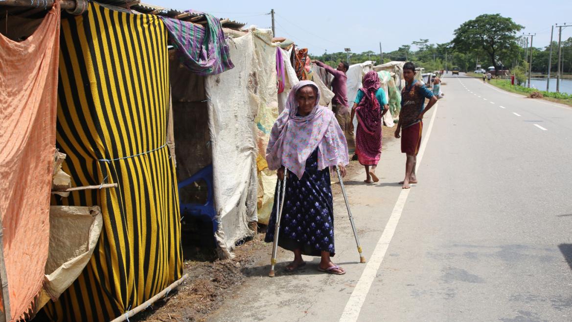 An older brown woman wearing a dress and head scarf walks alongside some makeshift dwellings with cloth covered entrances. She is using crutches, and walking alongside a highway.