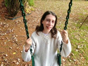 Foto de Sheryl Grossman sentada en un columpio. Es una mujer de tez clara y sonriente con cabello castaño hasta los hombros y un suéter blanco.