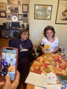 Anna and Judy in Judy's house, smiling at the camera. Anna holds a copy of Judy's memoir. Judy has her hand on Anna's arm.