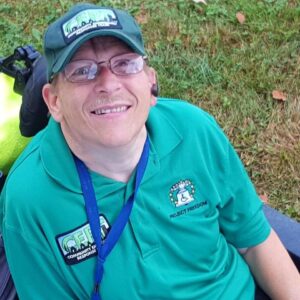 Norman Smith, a smiling white man wearing a green shirt and a baseball hat, seated in his wheelchair.