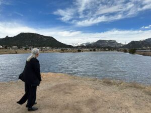 The image shows Melissa Marshall standing on the bank of a large body of water with mountains in the background. Melissa is wearing a black jacket and looking out over the water. The sky is clear and blue, with a few clouds visible in the distance. The mountains in the background are covered in snow and have trees growing on them. The water is calm and reflects the sky and mountains in its surface.