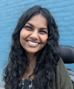 Priya Penner, a young brown woman with long, curly black hair and a nose piercing, smiles at the camera. She’s wearing a floral dress and a dark green cardigan.