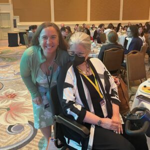 Melissa Marshall, an older white woman with COVID-gray hair sitting in a scooter, smiles next to Meg Traci, a white woman with brown hair.