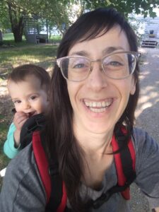 Photo of Kira Meskin smiling at the camera with her daughter, a toddler on her back.