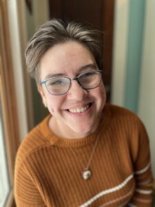 Photo of Jennith smiling at the camera. Jennith is a white person with short hair, and is wearing glasses and a burnt orange sweater.