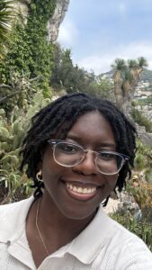 Image of Trinity, a dark-skinned Black femme with ear-to-neck length locs, purple and rose-gold glasses, small hoop gold earrings, a gold necklace, and wearing a cream-colored blouse with a collar. She is smiling in front of cacti and other native plants in Ezé, France underneath the blue, cloudy sky.