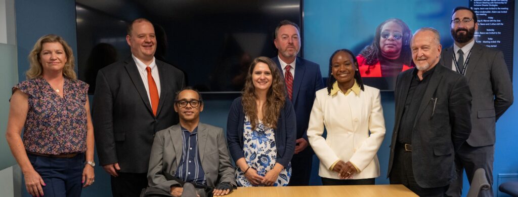 Nine people pose together in a conference room following a meeting between The Partnership for Inclusive Disaster Strategies and FEMA. Eight people are in the room, including one wheelchair user seated in front, while another participant appears virtually on a large screen behind the group. A conference table is visible in the foreground. [Photo credit: FEMA]